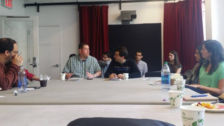 A group of people sit around a white table in a conference room. Two men having a conversation sit at the head of the table. 