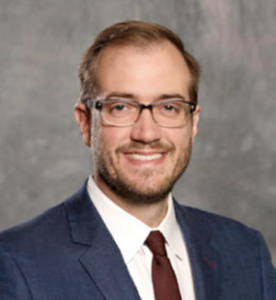 A professional portrait in which a man with square glasses smiles at the camera.