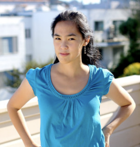 A photo of a young East Asian woman with long hair and wearing a blue top. She is standing outside in front of a row of white buildings.