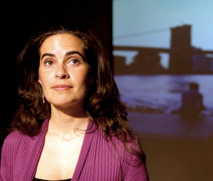 A photo of a woman with light skin and long brown hair wearing a mulberry sweater. She is standing in front of a projected image of what appear to be buildings.
