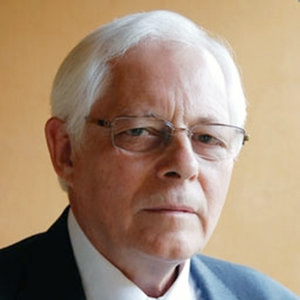 a headshot of an unsmiling white man with thinning white hair and rectangular glasses.