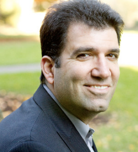 A three-quarter portrait of a man with very short brown hair and a dimpled chin. He is wearing a suit.