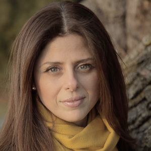 A light skinned woman with light eyes and medium brown, straight hair wears a yellow scarf and looks into the camera. 