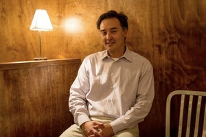 A portrait of a man wearing a white shirt and cream pants sitting against a wooden wall. A lamp next to his head casts the entire photograph in a yellow glow.