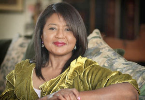 A woman with medium-brown skin, straight brown hair, red lipstick, and a light green velvet shirt smiles at the camera