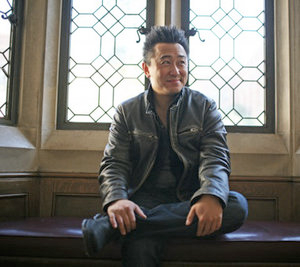 A photo of an East Asian man with spiky hair and wearing a leather jacket, sitting with one leg crossed over the other in front of a set of ornate windows.