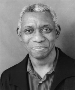 A grayscale portrait of a black man with salt and pepper hair and wearing a dark jacket smiling at the camera.