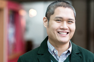 An Asian man with a salt-and-pepper buzz cut and wearing a forest green coat smiles into the camera