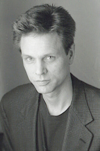 A thin, light skinned man in T-shirt and jacket looks into the camera.
