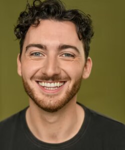 A young man with a short brown beard and very short curly hair smiles into the camera.