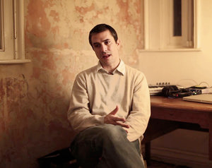 A seated man in a cream colored sweater converses with someone behind the camera.