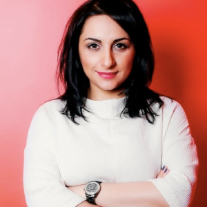 A dark-haired young woman in a white sweater stands with arms crossed in front of a red background.