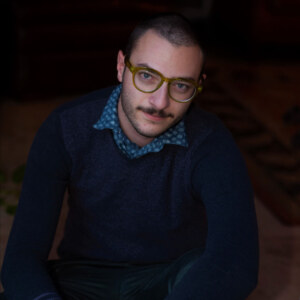 A young man wearing brown glasses and a dark blue sweater looks into the camera.