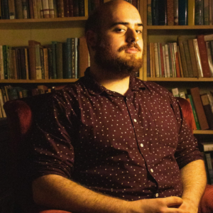 A bald man with a dark beard and mustache wearing a dark red shirt looks at the camera.