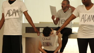Four people in white V-neck T-shirts stand three in front, one in back. Of the three in front, two are labeled Adam, one is labeled Eden. The Adams are on either side of Eden, who is kneeling. The person in the back is labeled Eve.