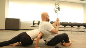 A profile view of a man in a white T-shirt sitting on the floor, holding a branch in front of his face. He leans against the shoulder of another person in black pants and white shirt.