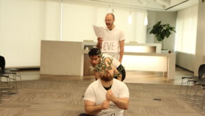 Three people in white T-shirts stand in a line. The person in the back reads from a script. The person in the front holds a branch in front of his face. The third person crouches between them.