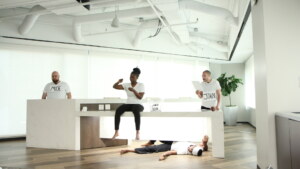 three people in white T-shirts are at a white table. Two stand at either end, while one sits on the table in the middle.