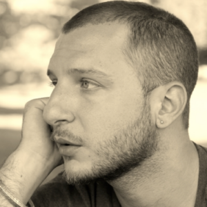 A young man in profile, looking to the left. He has a shaved head and short beard and is wearing a tiny gold earring.