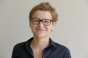 A person with very short dark blonde hair and black rectangular glasses smiles at the camera