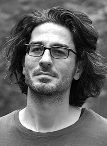 a light-skinned man with a scruffy beard, shoulder-length hair, and dark rectangular glasses looks slightly to the left of the camera.