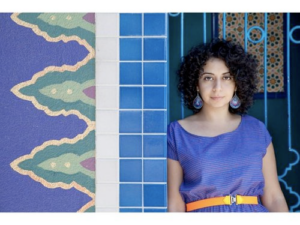 A woman with dark curly hair wearing big blue earrings, blue dress, and yellow belt leans against a blue tiled wall.