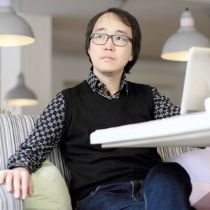 A seated, serious man in a black sweater vest, dark jeans, and dark glasses looks to the left of the camera.
