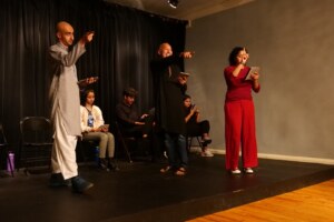 Three people stand on a stage and point at the audience. The one on the left is wearing gray, the one in the middle black, and the one on the right bright red.
