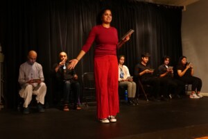 a woman in a bright red outfit stands on a stage. Six people in gray and black sit behind her.