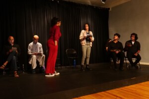 Six people stand or sit on a stage. Two women converse in the middle. The one on the left is wearing a cherry red shirt and pants. The one on the right wears cream and light green.