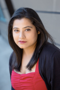 A young woman with straight dark hair and wearing a red tank top and black sweater looks into the camera.