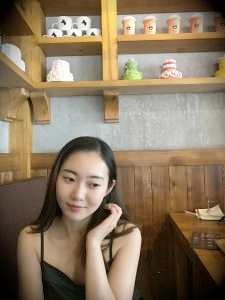 An East Asian woman in a spaghetti strap tank top looks down and to the right. One hand is tucking her hair behind her ear.
