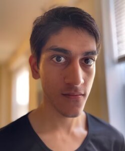 A young South Asian man with very short brown hair looks seriously into the camera.