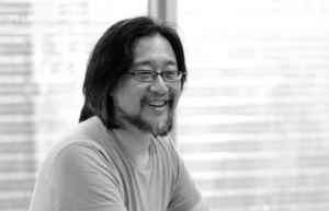 A black and white portrait of an East Asian man with shoulder length hair, a goatee, and rectangular glasses.