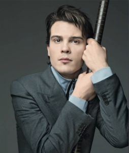 A portrait of a pale, serious-looking young man with short brown hair and wearing a gray suit holding on to a pole.