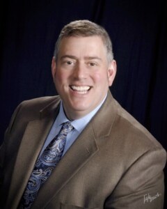 A man in a brown suit smiles at the camera