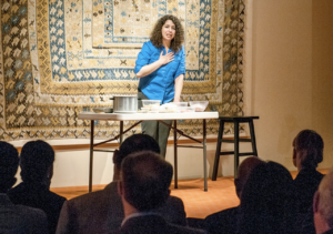 A woman with very curly hair and in a blue top stands behind a card table. She is speaking passionately to an audience. Behind her is a large blue and ivory patterned hanging artwork.